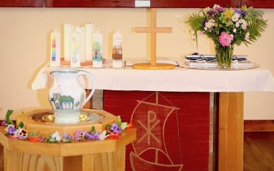 Auf dem Bild sieht man den Altar, im Vordergrund ist das Taufbecken zu sehen. Auf dem Altar stehen Blumen, Kerzen und ein helles Holzkreuz.