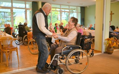 Auf dem Bild sieht man einen älteren Herren mit einer älteren Dame tanzen. Die Frau sitzt im Rollstuhl.