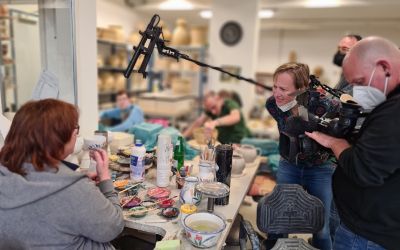 Auf dem Bild sieht Reporterin Theresa Majerowitsch, wie sie einer Keramikmitarbeiterin fragen stellt. Neben ihr sieht man den Kameramann und den Mikrofonmitarbeiter. Auf dem Tisch stehen Farbtöpfe und Pinsel.
